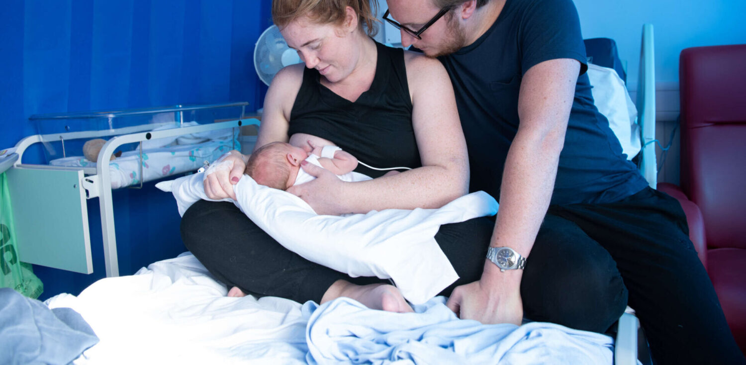 A mother breastfeeds her newborn in the maternity unit alongside the father