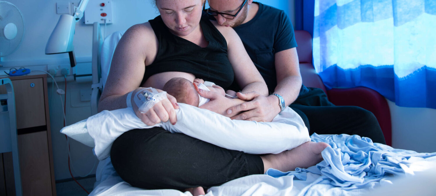 A mother breastfeeds her newborn in the maternity unit alongside the father