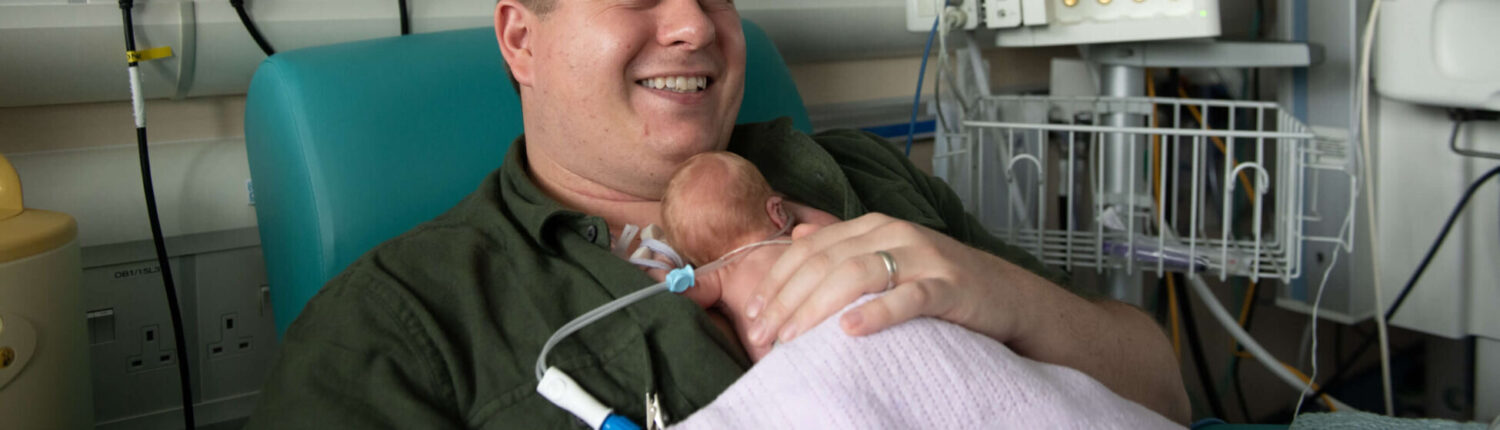 A father of twins born premature holds his infant in skin-to-skin contact on the neonatal unit.