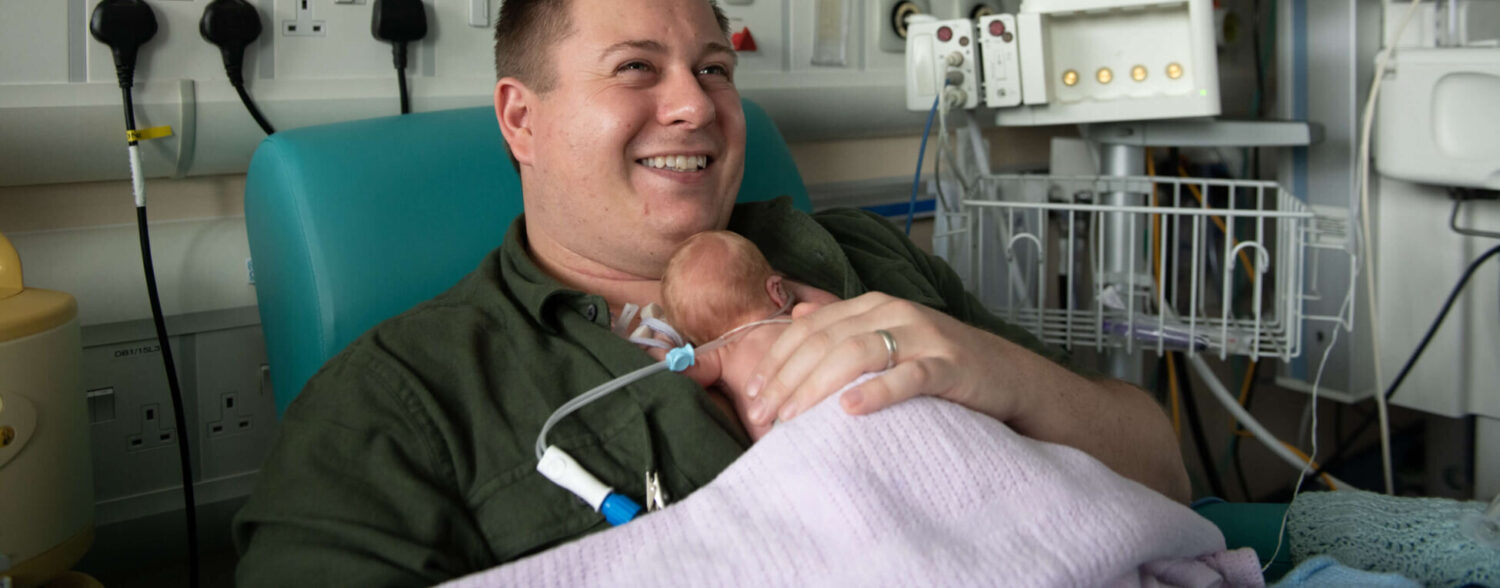 A father of twins born premature holds his infant in skin-to-skin contact on the neonatal unit.