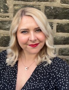 A smiling blonde woman is seen from the chest up in front of a stone wall. She wears a navy polka dot dress and a gold pendant necklace.