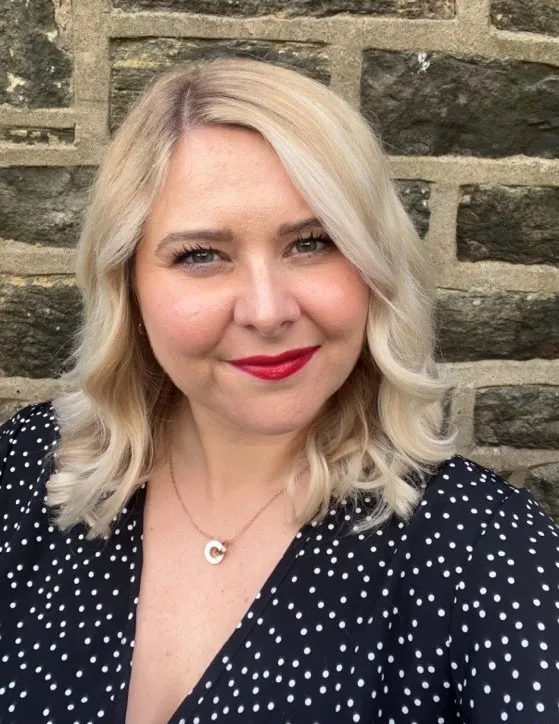 A smiling blonde woman is seen from the chest up in front of a stone wall. She wears a navy polka dot dress and a gold pendant necklace.