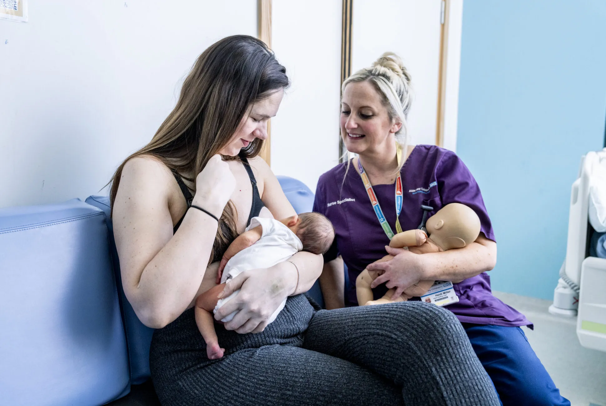 Mother Jen, with long brown hair and a black nursing top, and midwife Heidi, a smiling blond woman in purple scrubs, check baby Emilia's latch as Jen sits to feed Emilia at Alder Hey hospital in Liverpool.