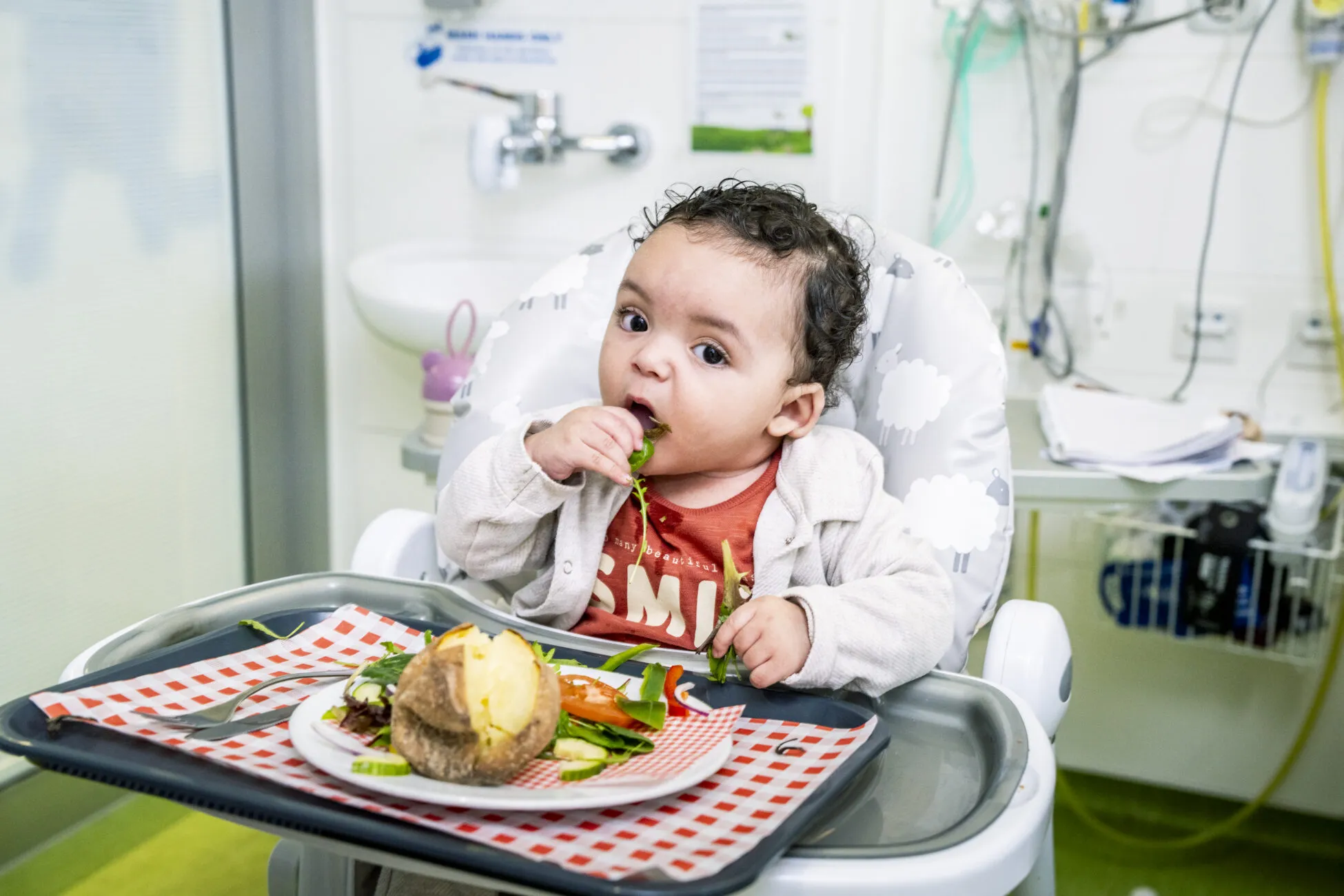 Baby Maalik brings salad to his mouth as he sits in his high chair at Alder Hey hospital. In front of him is a plate with a jacket potato and vegetables on a red and white check tray.