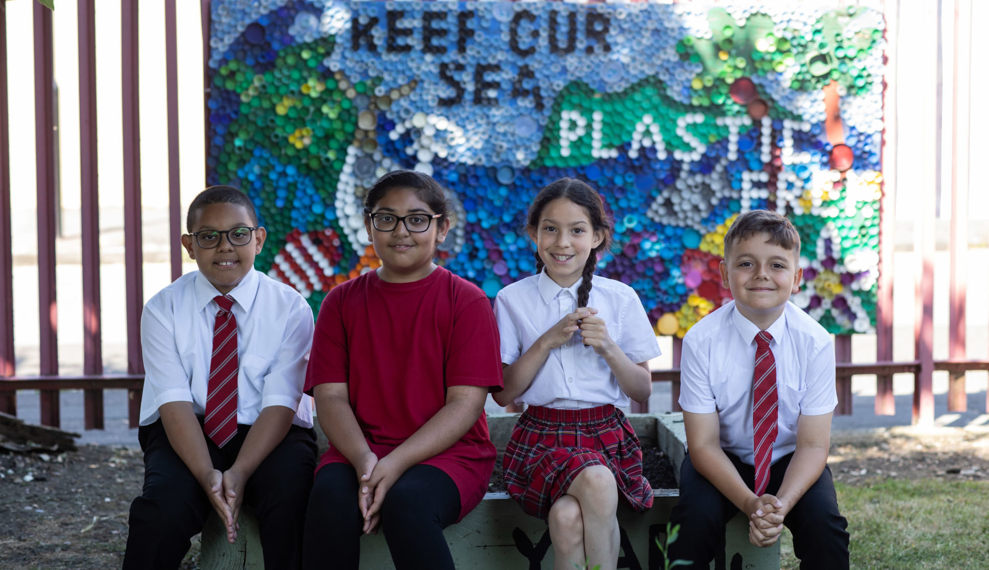 A group of four children posing in front of a 'Keep our see plastic free' display they created out of plastic lids