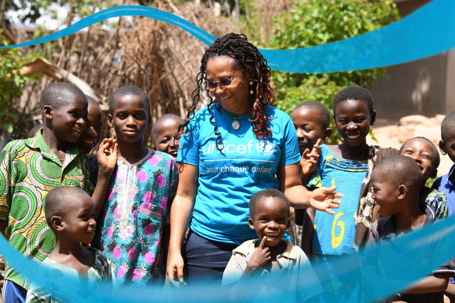 Image shows a UNICEF staff memeber standing with smiling children from a country office. The staff member is wearing a cyan blue and white UNICEF t-shirt.