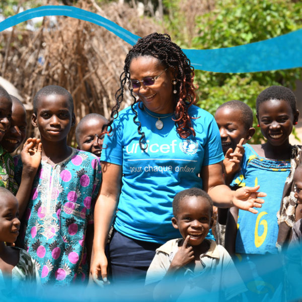 Image shows a UNICEF staff memeber standing with smiling children from a country office. The staff member is wearing a cyan blue and white UNICEF t-shirt.