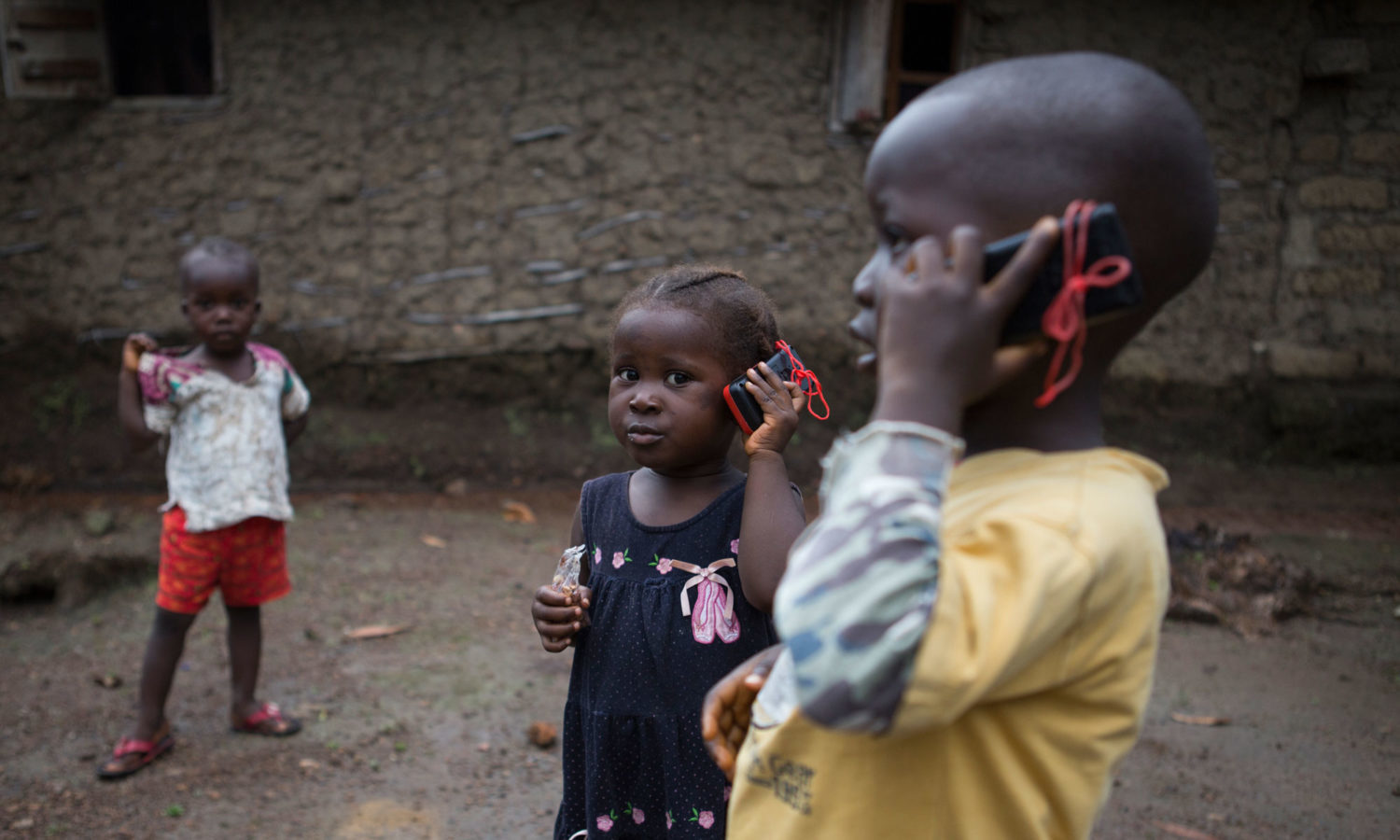 On 25 September, [NAMES CHANGED] (foreground, left-right) siblings Awa, 3, and Amadou, 4, pretend to talk on toy mobile telephones, at home in a village on the outskirts of the city of Kenema in Eastern Province. Many of the people in the village are affected by Ebola. Olematu and Ibrahim are being cared for by their 15-year-old sister, Betty, with help from a neighbour, following the death of their mother from Ebola one month ago. Betty is a survivor of the virus. An estimated 1,131 children in Sierra Leone are affected by Ebola virus disease. UNICEF/UNI172461/Bindra