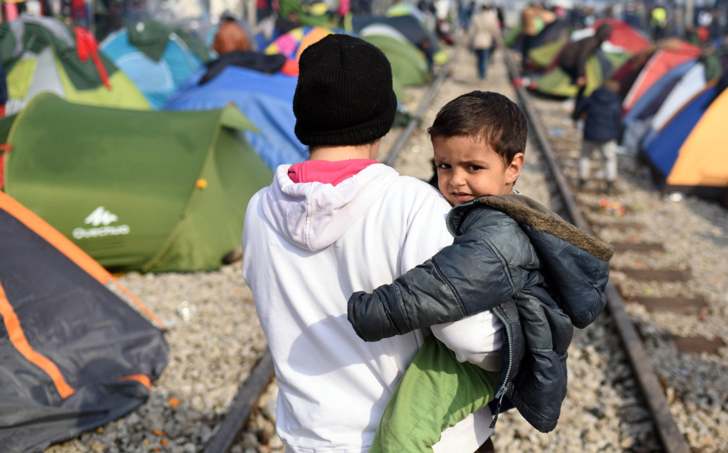A young boy is carried by a family member walking between makeshift tents along the train tracks in Idomeni, Greece. Unicef/2016/Georgiev