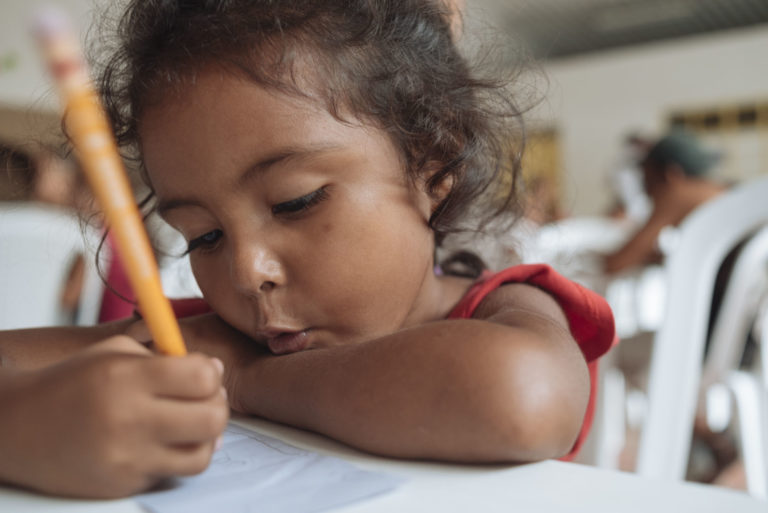 Young girl at a desk drawing a picture with a pencil.