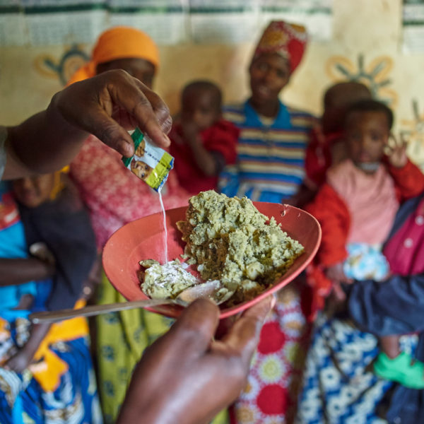 A community health worker adds micronutrient powder to a child's food, as children and their parents wait in the background.