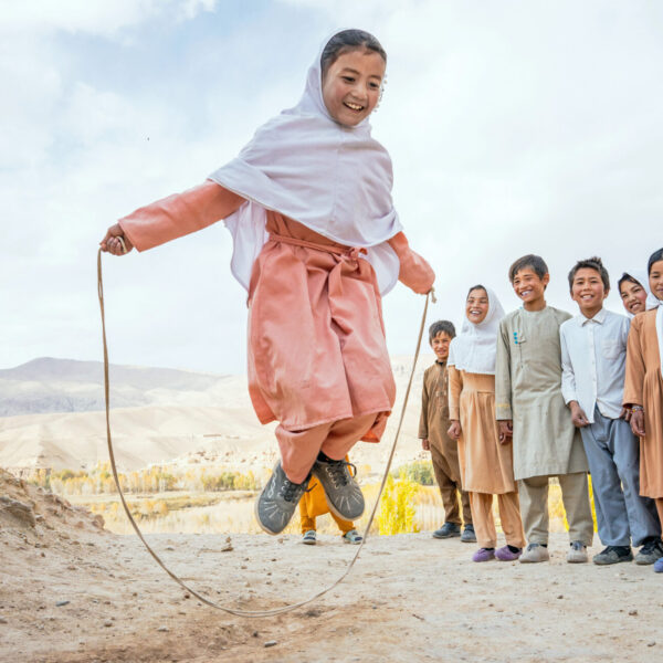 Girl in a pink dress and white headscarf smiles as she plays with skipping rope while other children watch glee.
