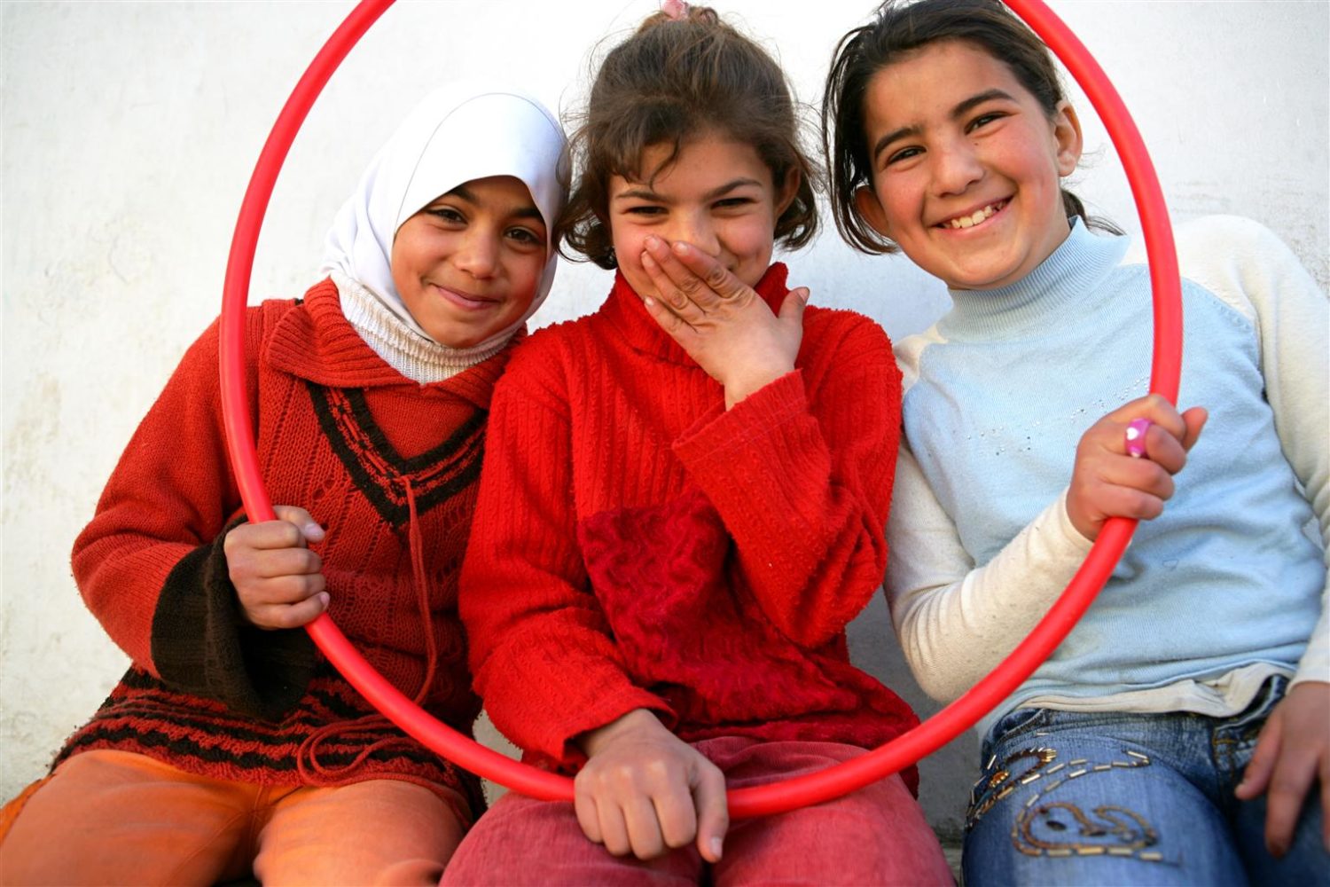 3 girls sitting down holding up a hula hoop and smiling.