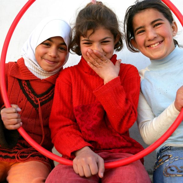 3 girls sitting down holding up a hula hoop and smiling.