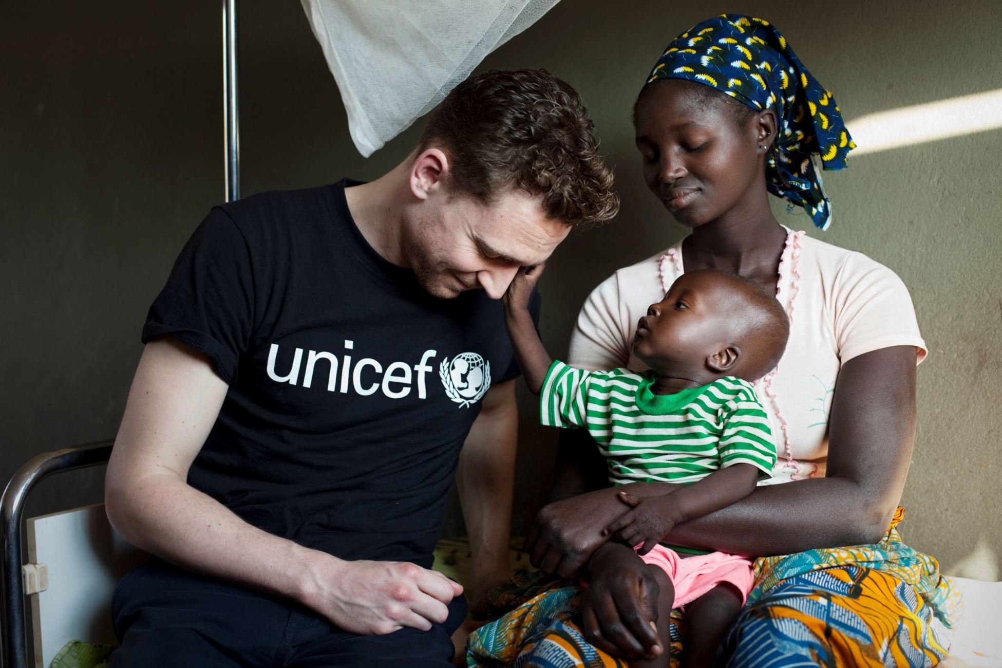 A baby, sitting on his mother's lap, with Tom Hiddleston at the National Institute of Child Health and Nutrition in Guinea