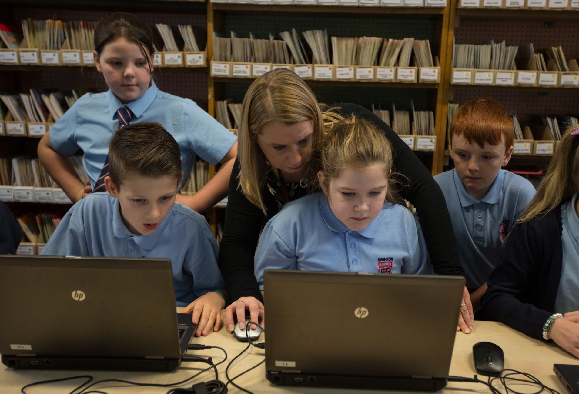 A group of children looking at the screens of two laptops with a woman supervising.