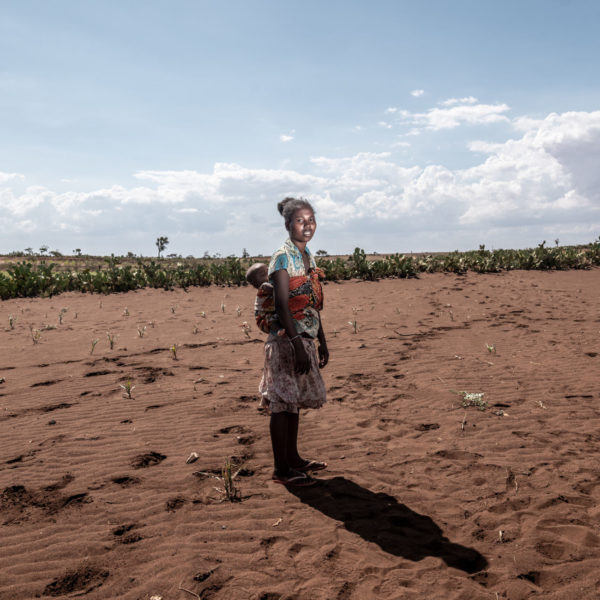 A mother with her baby strapped to her back stands in her family's empty corn field.