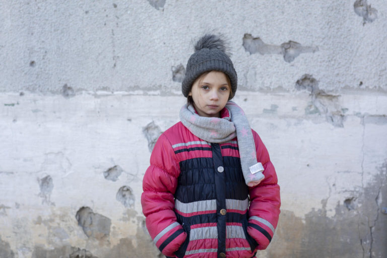 On 13 February 2022, 9-year-old Masha stands in front of the conflict-damaged exterior of her home in eastern Ukraine.