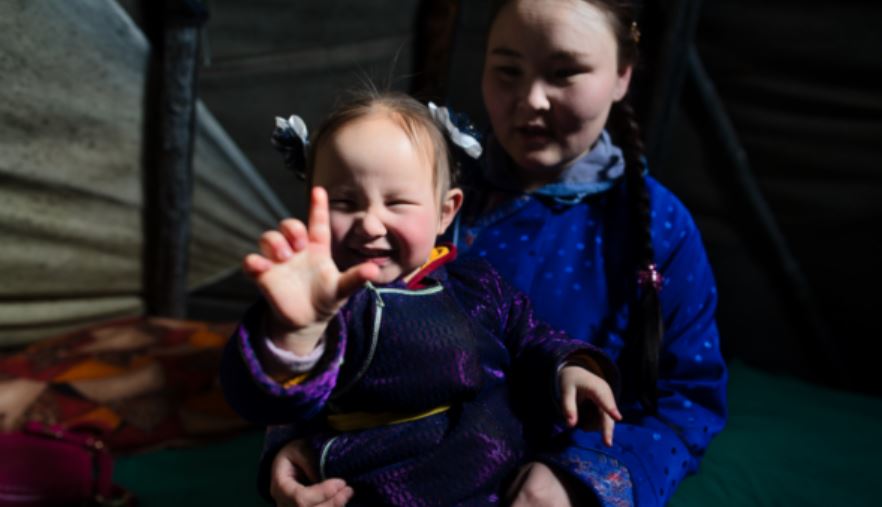 Anirlan in Mongolia holds up her inked finger to the camera, indicating that she has recently been vaccinated. Her mum holds her in the background.