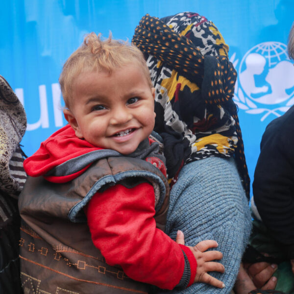 A child smiles while queuing with his mother in front of a UNICEF health and nutrition mobile clinic for consultation and distribution of medicine at Musab Bin Omair mosque, Tal Hajar, Hasakah city, northeast Syrian Arab Republic, on 27 January 2022. The mosque is currently a temporary shelter for some 120 internally displaced families. Some 45,000 people were forced to flee their homes due to conflict. As violence continues in and around the Ghwayran/ Sinai’i detention centre, in northeast Syria, children continue to be critically vulnerable and in urgent need of protection. Violence forced thousands of people in the area to flee, most are women and children. Some have been displaced several times fleeing violence in other parts of Syria over the years. UNICEF is on the ground, working with partners, to provide displaced children and their families with lifesaving assistance, including clean water and critical hygiene supplies. UNICEF’s volunteers have helped people reach shelters and clinics and distributed food, blankets, mattresses, clothes, and medicine. They have also distributed materials on the risks of explosive ordnance to raise awareness among boys and girls in the shelters and keep them safe. A UNICEF-supported mobile health and nutrition team is providing services and medicine to vulnerable children and their mothers. To date, the team has provided health consultations and free medicine; screened children, pregnant and lactating women; and provided malnourished children with ready-to-use supplementary food. UNICEF is providing information to families on how to prevent separation and access psychosocial support to children and caregivers.