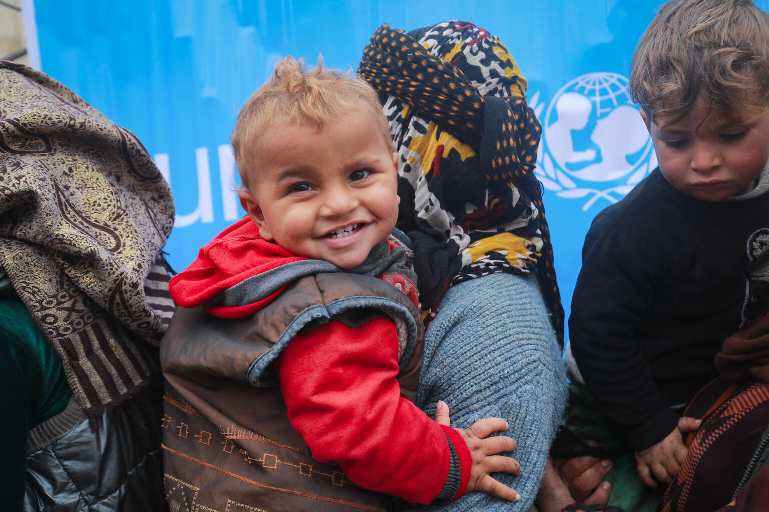 A child smiles while queuing with his mother in front of a UNICEF health and nutrition mobile clinic for consultation and distribution of medicine at Musab Bin Omair mosque, Tal Hajar, Hasakah city, northeast Syrian Arab Republic, on 27 January 2022. The mosque is currently a temporary shelter for some 120 internally displaced families. Some 45,000 people were forced to flee their homes due to conflict. As violence continues in and around the Ghwayran/ Sinai’i detention centre, in northeast Syria, children continue to be critically vulnerable and in urgent need of protection. Violence forced thousands of people in the area to flee, most are women and children. Some have been displaced several times fleeing violence in other parts of Syria over the years. UNICEF is on the ground, working with partners, to provide displaced children and their families with lifesaving assistance, including clean water and critical hygiene supplies. UNICEF’s volunteers have helped people reach shelters and clinics and distributed food, blankets, mattresses, clothes, and medicine. They have also distributed materials on the risks of explosive ordnance to raise awareness among boys and girls in the shelters and keep them safe. A UNICEF-supported mobile health and nutrition team is providing services and medicine to vulnerable children and their mothers. To date, the team has provided health consultations and free medicine; screened children, pregnant and lactating women; and provided malnourished children with ready-to-use supplementary food. UNICEF is providing information to families on how to prevent separation and access psychosocial support to children and caregivers.