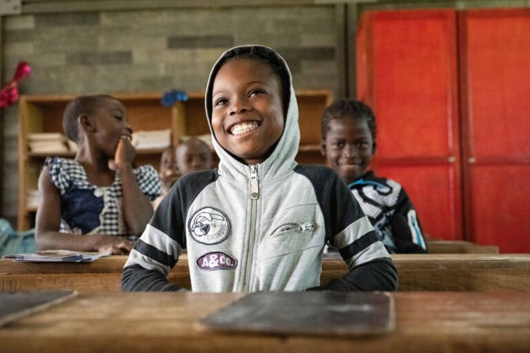 A girl sits behind her desk and smiles among her classmates.