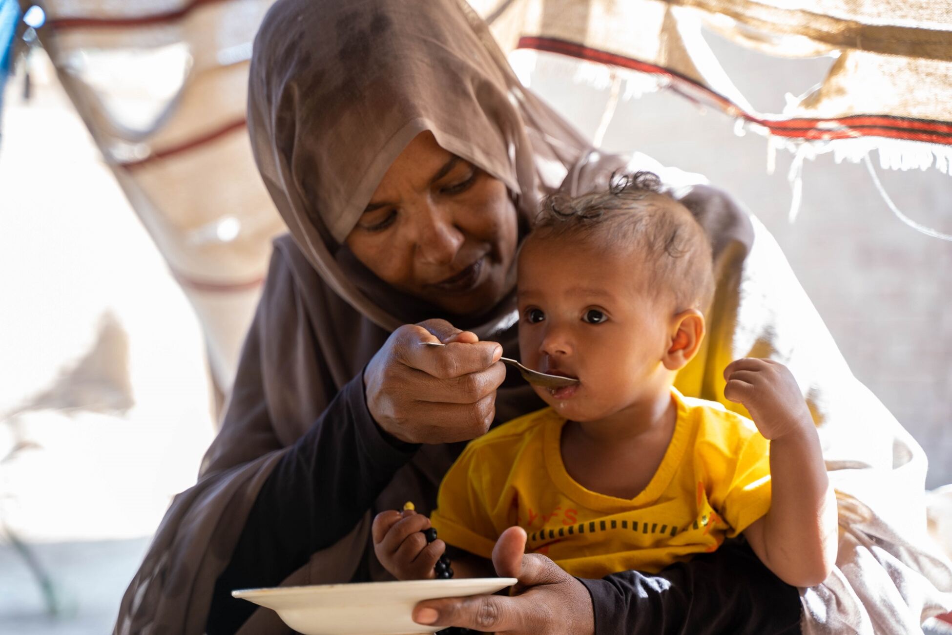 A mother feeds lentils to her daughter who is sitting on her lap.