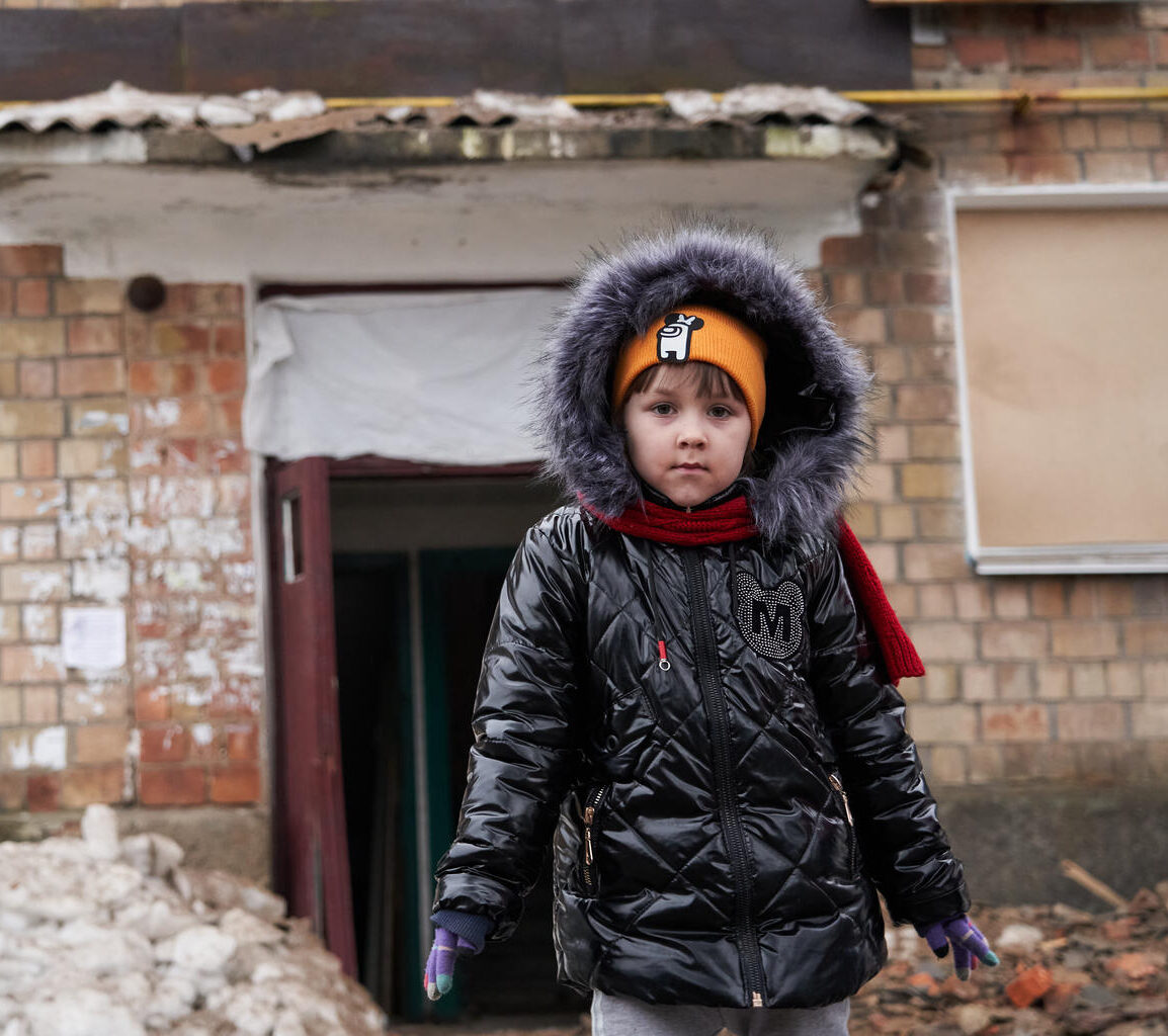 A young girl bundled up in winter clothing standing in front of a dilapidated building.