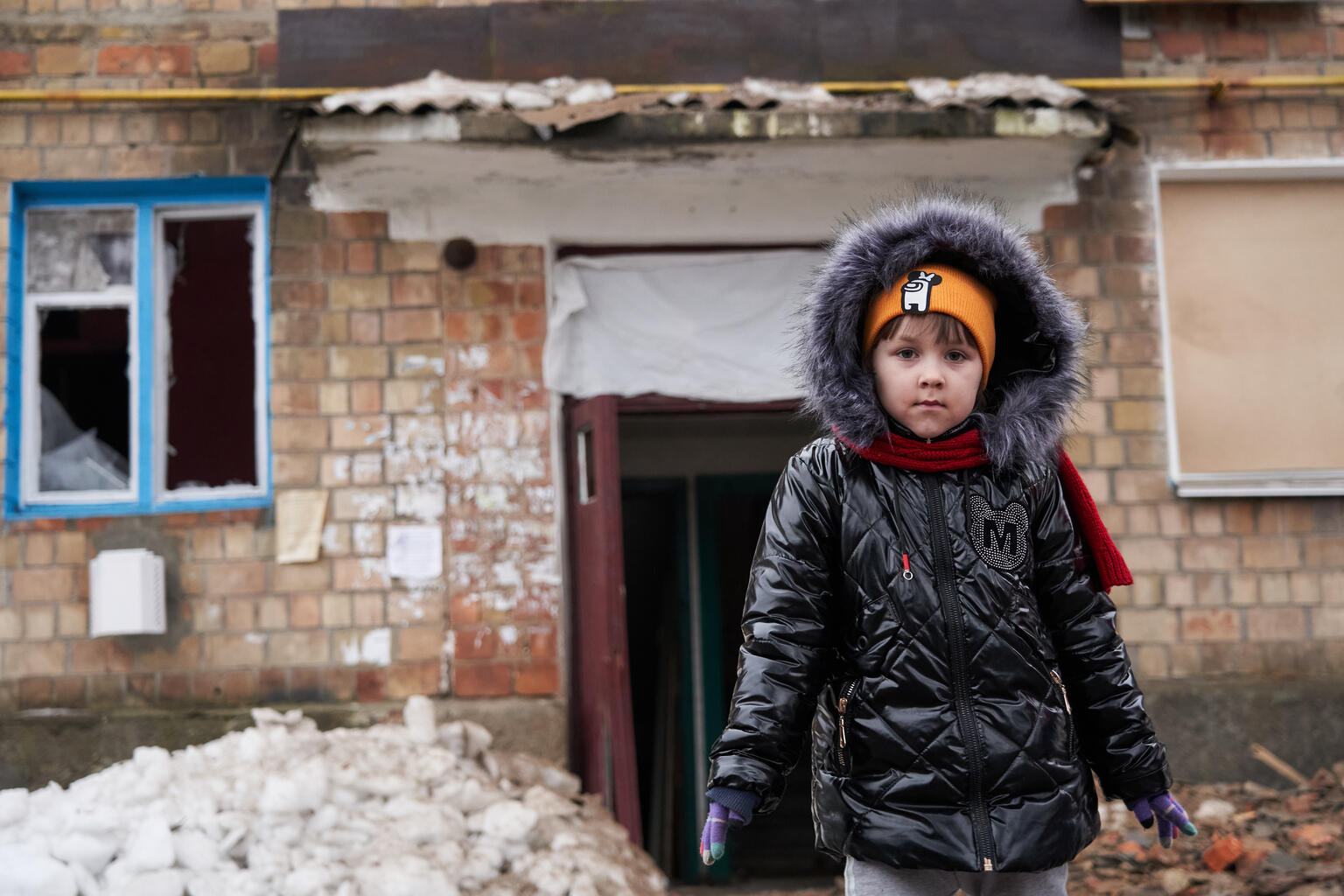 A young girl bundled up in winter clothing standing in front of a dilapidated building.