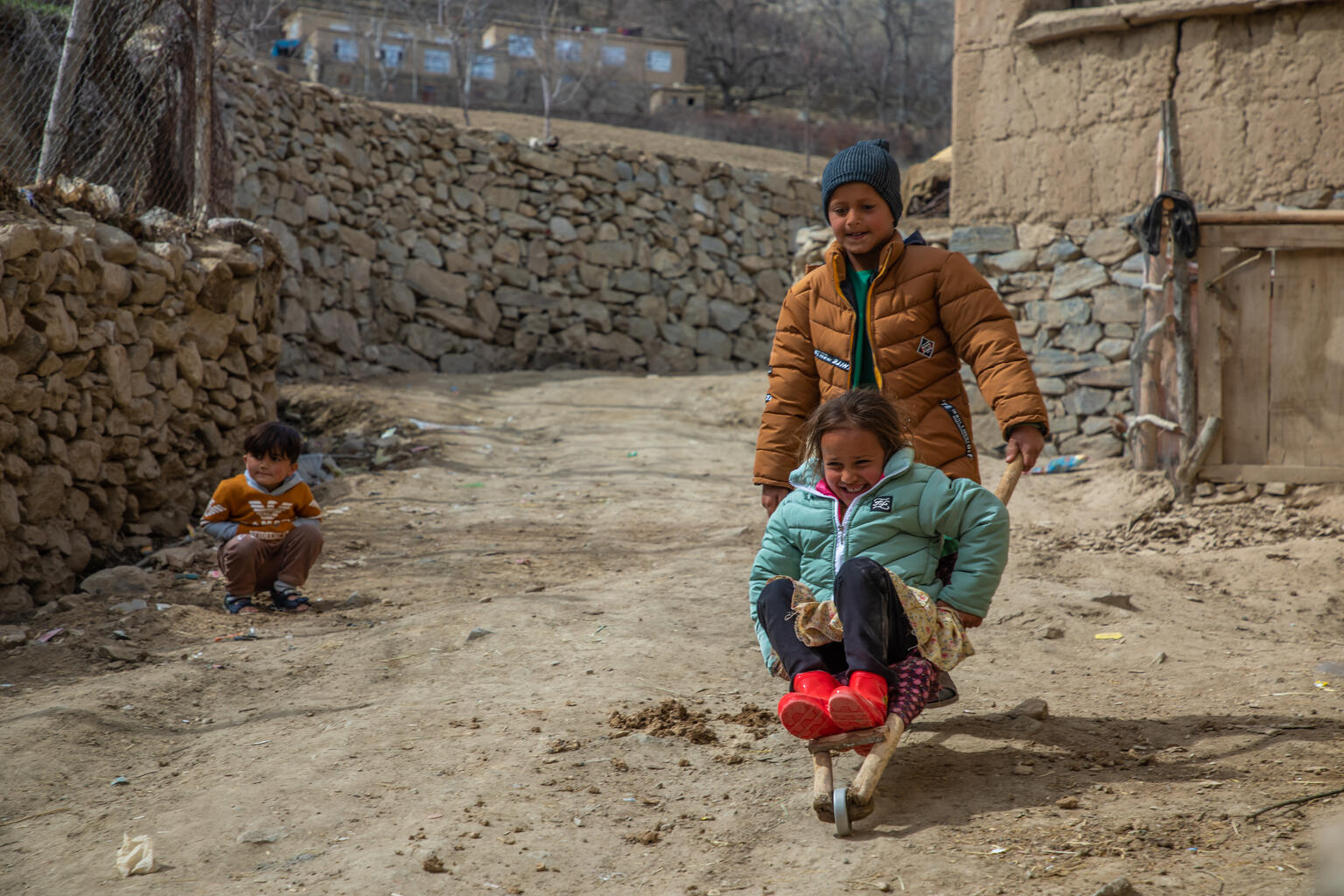 an older boy pushes a younger girl on a trolley, both are smiling.
