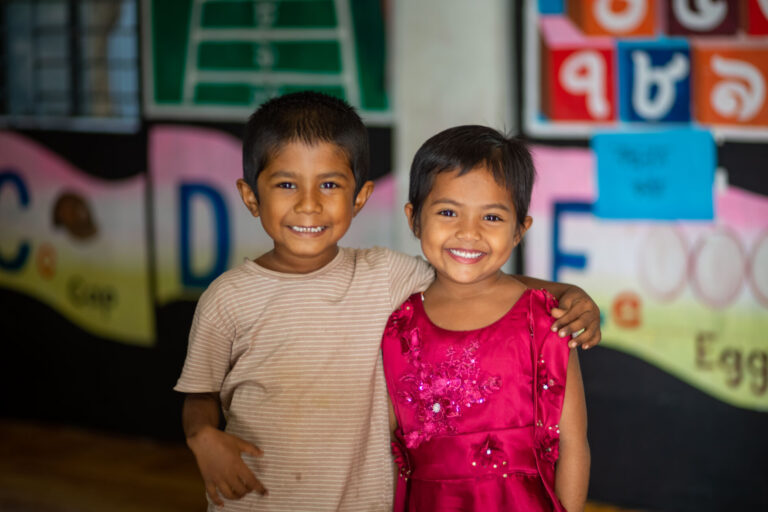 Two girls from a UNICEF-funded day care center in Bangladesh smile at the camera with their arms around each other
