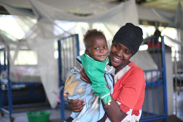 A mother holds her smiling baby on a hopsital ward. Mosquito nets are in the background.