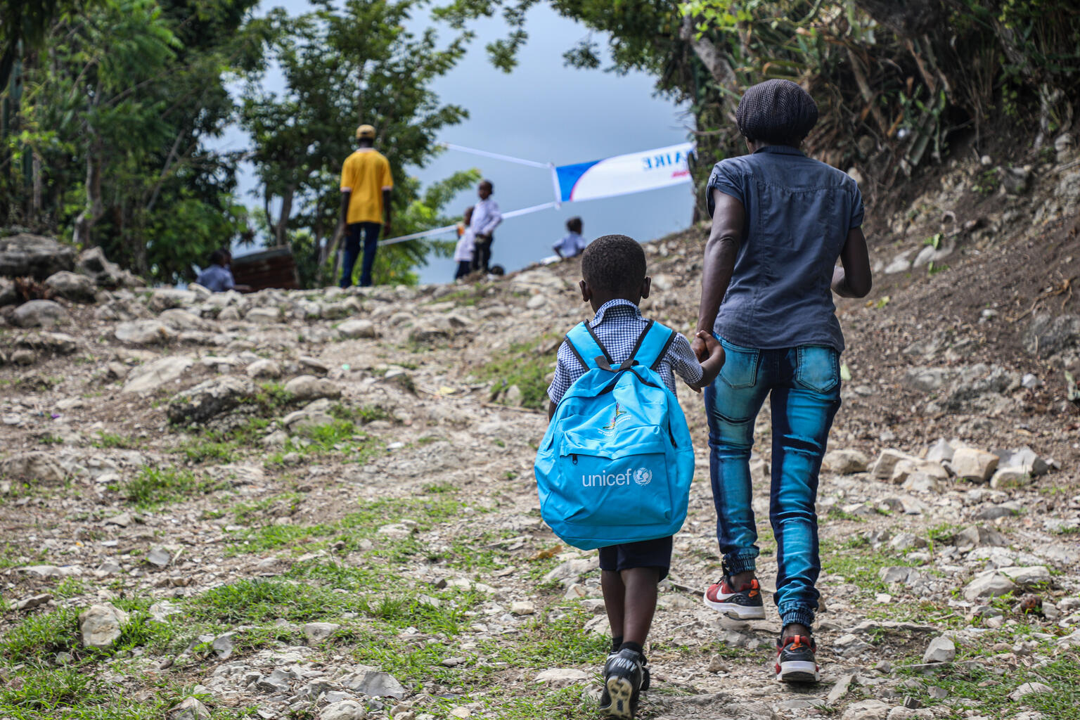 A mother and child walk hand-in-hand in Haiti. The child is wearing a blue unicef backpack.