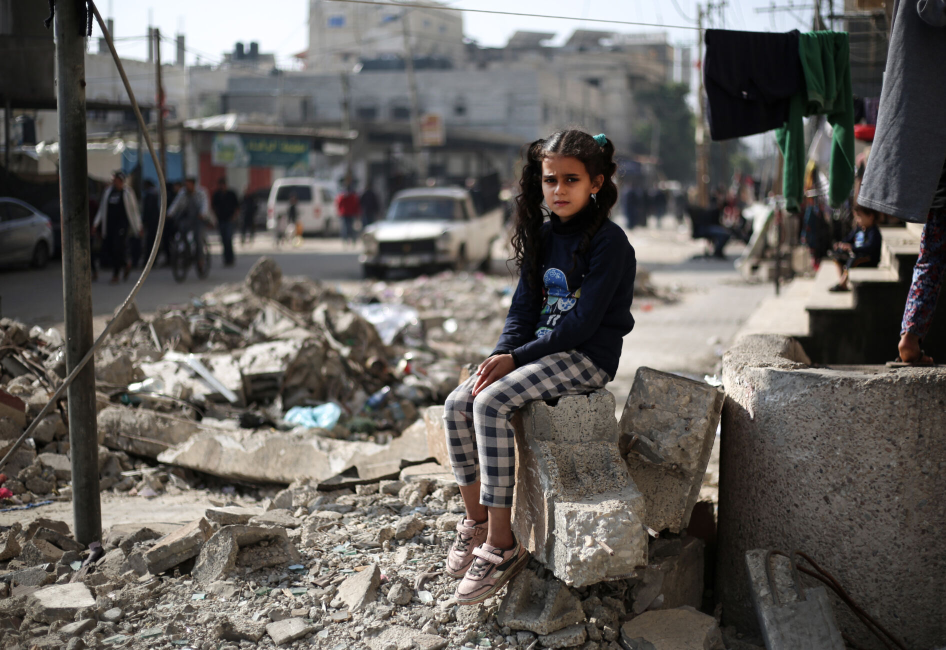 A girl in a black jumper and black and white trousers sits on a piece of rubble in Rafah, south of Gaza.