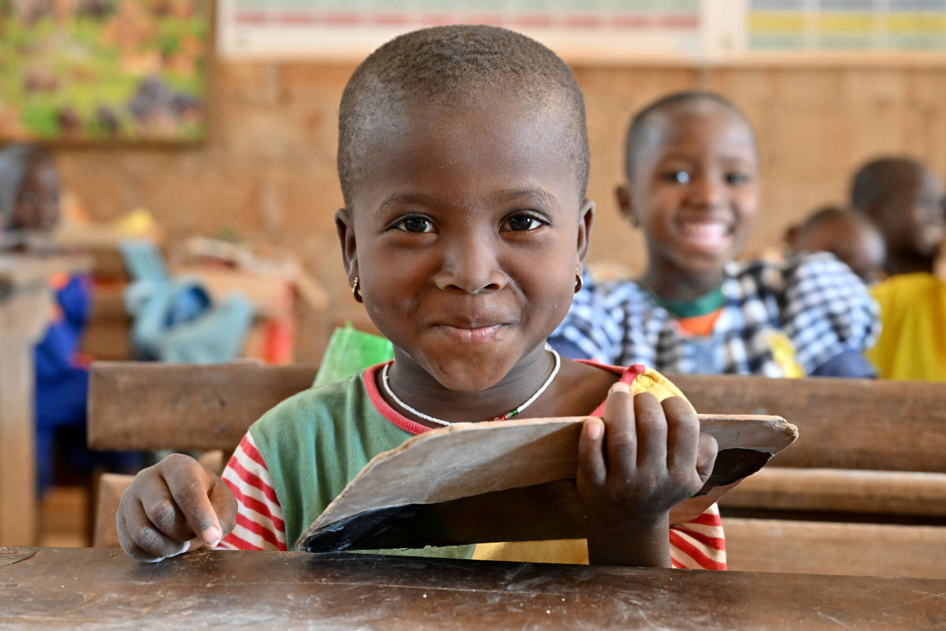 A young child looks up from her work and smiles at the camera from behind a wooden schooldesk.