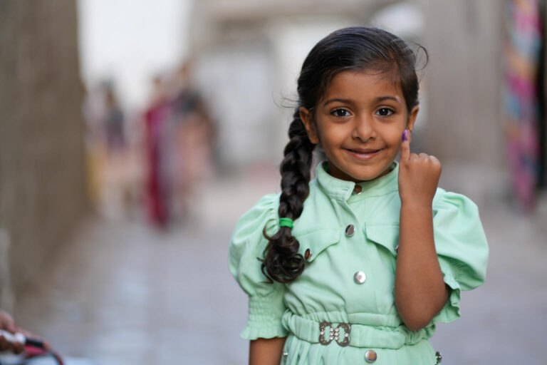 A young girl smiles and holds up her little finger, which has been painted purple to indicate she has been vaccinated.