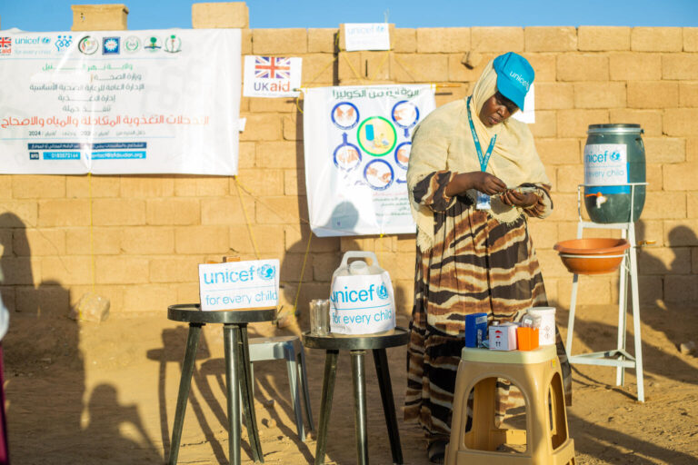 A UNICEF volunteer prepares to provide WASH and nutrition services to children and mothers beside an information table and a water container.