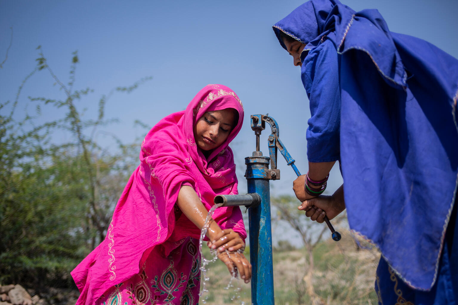 Two girls in brightly coloured clothing pump clean water in Pakistan.