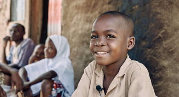 8-year-old Abasse sits and smiles outside his home, his family in the background.