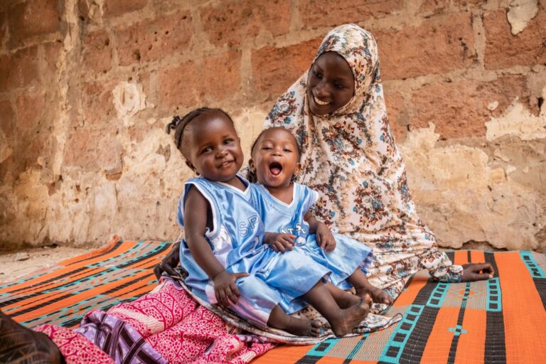 The twins Asma'u and Halima sit on the carpet. Halima is smiling and Asma'u is making a silly face. Their mother looks at them smiling.