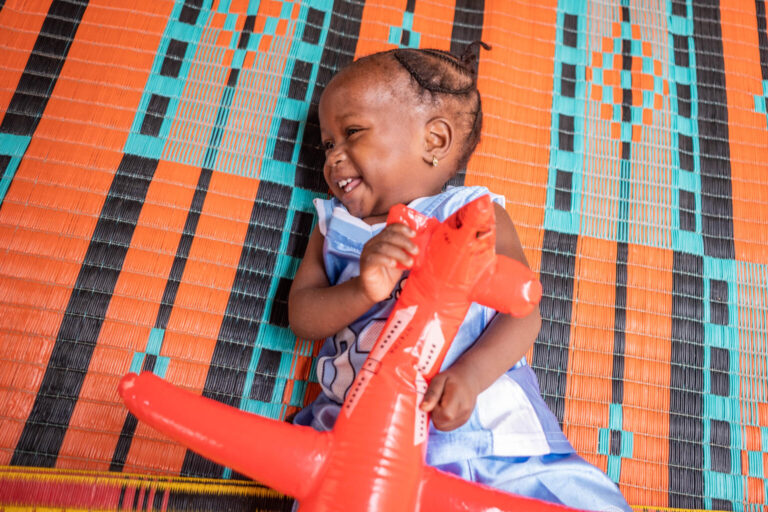 14-month old Asma'u plays with an inflatable aeroplane and giggles on the carpet.
