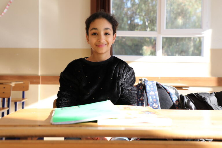 14-year-old Ela sits behind her school desk smiling.