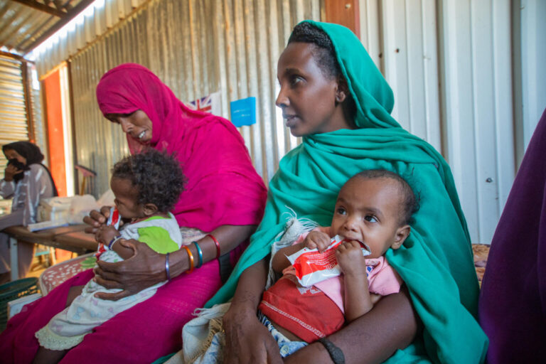 A mother holds her two-year-old across her lap at a nutrition screening. Her child is eating therapeutic food from a red package.