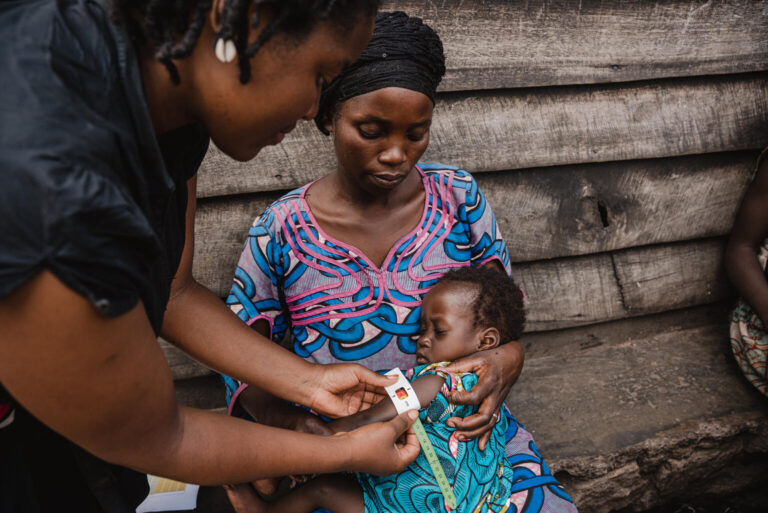 A mother holds her young child tightly as a health worker measures the child's upper arm as part of a nutrition screening.