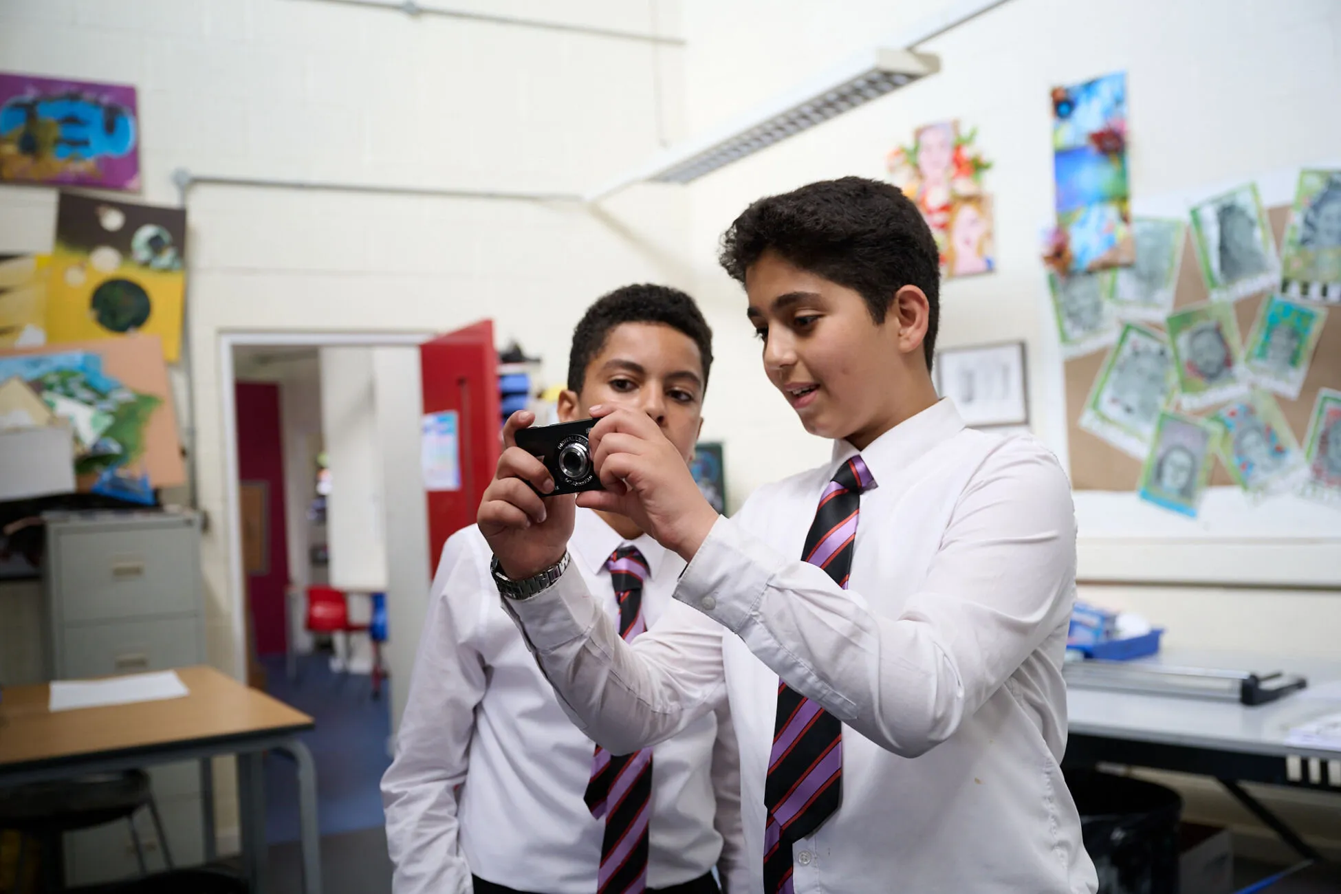 There are two schoolboys in a classroom, and one of them is holding a small camera.