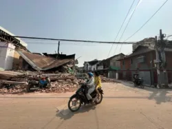 Two people travel by motorbike past the remains of collapsed buildings.