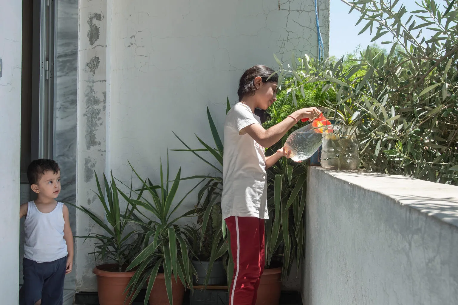 Lana, 12, Leila’s granddaughter, waters the plants on her balcony in Western gathering, Kisweh city, Rural Damascus, Syria, while a younger grandchild looks on.