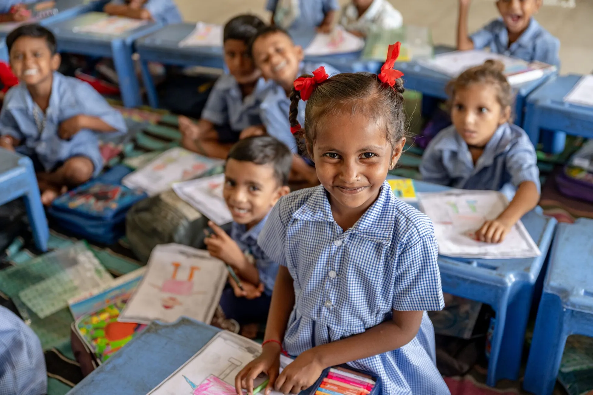 A 5 year old child in a primary school classroom, smiling while surrounded by classmates engaged in a colouring activity.
