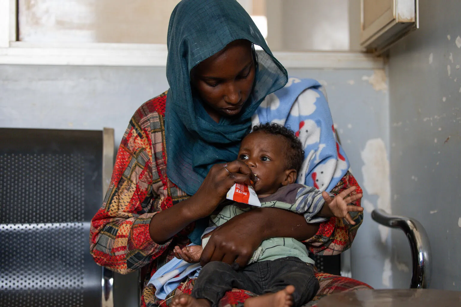 Naba holds her baby in her arms while feeding him from a red and white sachet of therapeutic food at a nutrition sight in Khartoum, Sudan,