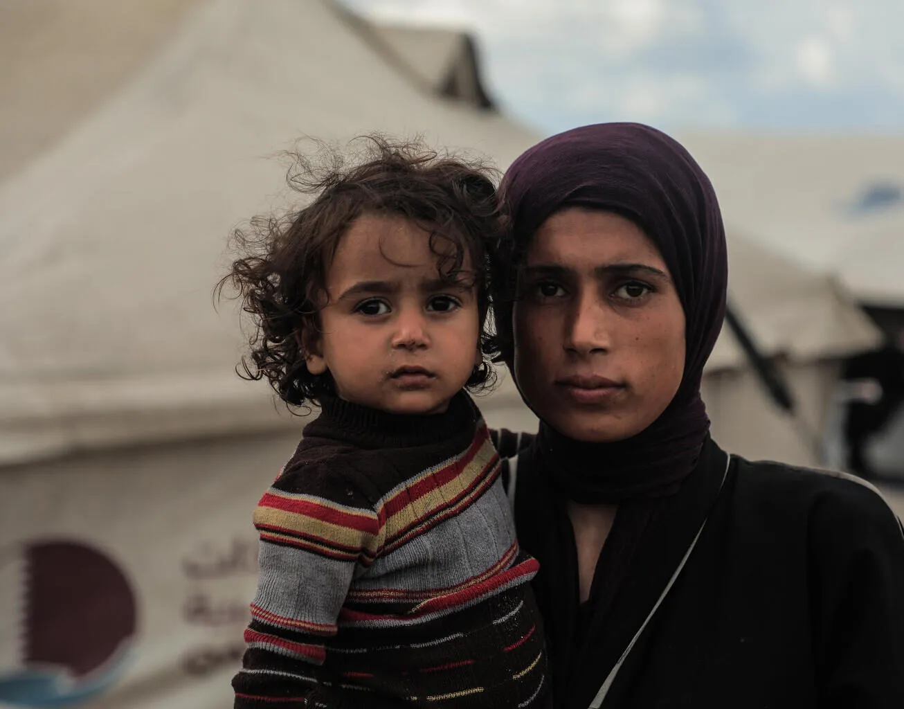 Maysaa, a mother of 5, looks solemn while holding her daughter outside their makeshift tent.